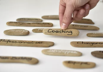 Main choisissant un galet en bois flotté portant l’inscription Coaching, parmi d’autres mots liés aux RH